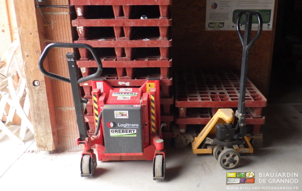 photo de basculeur à gauche et transpalette à droite rangés sous des piles de palettes